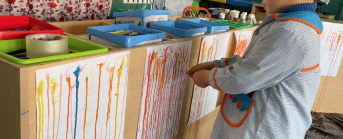 Niño de 4 años pintando de color naranja con un cuentagotas sobre un folio blanco colgado en vertical en un aula de educación infantil