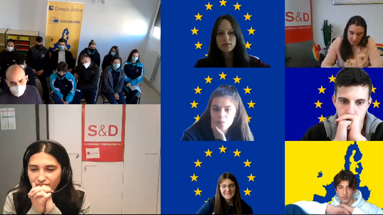 Captura de pantalla de una videoconferencia entre una eurodiputada y diversos jóvenes, algunos desde sus hogares con banderas de Europa y mapas como fondo, y otros en grupo desde clase sentados en sillas con mascarilla puesta