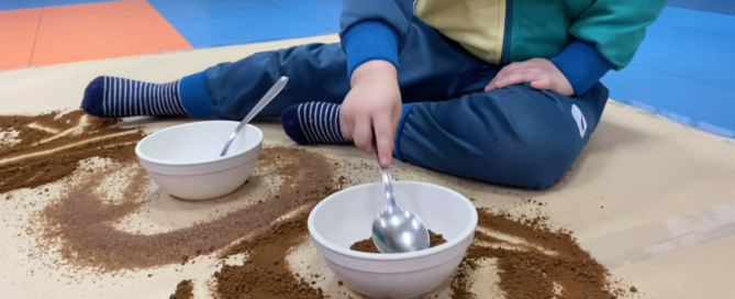 Niño de tres años sentado en el suelo de su clase jugando con una cuchara y cuencos con café y azúcar moreno