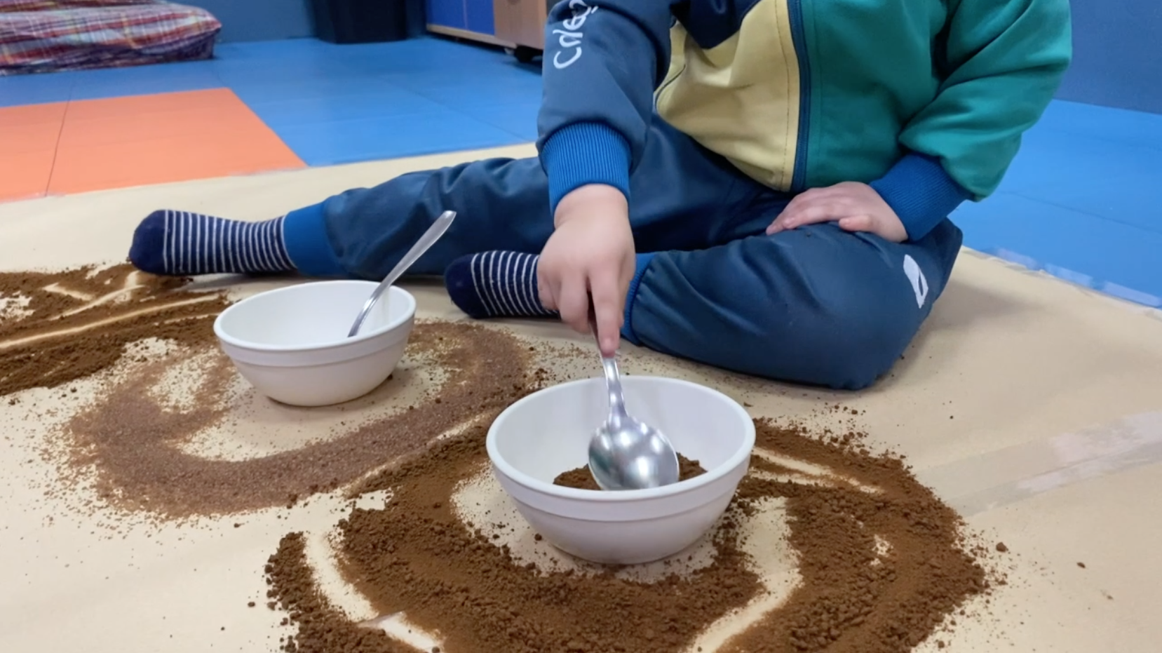 Niño de tres años sentado en el suelo de su clase jugando con una cuchara y cuencos con café y azúcar moreno